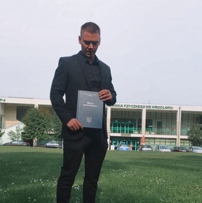 Trener personalny Wrocław z dyplomem AWF trzymający pracę magisterską przed Akademią Wychowania Fizycznego we Wrocławiu